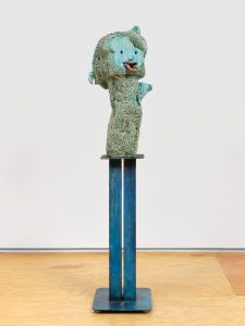 Turquoise-glazed ceramic figure with a doll-like face, textured surface, and small bird on top, mounted on a tall blue metal pedestal.