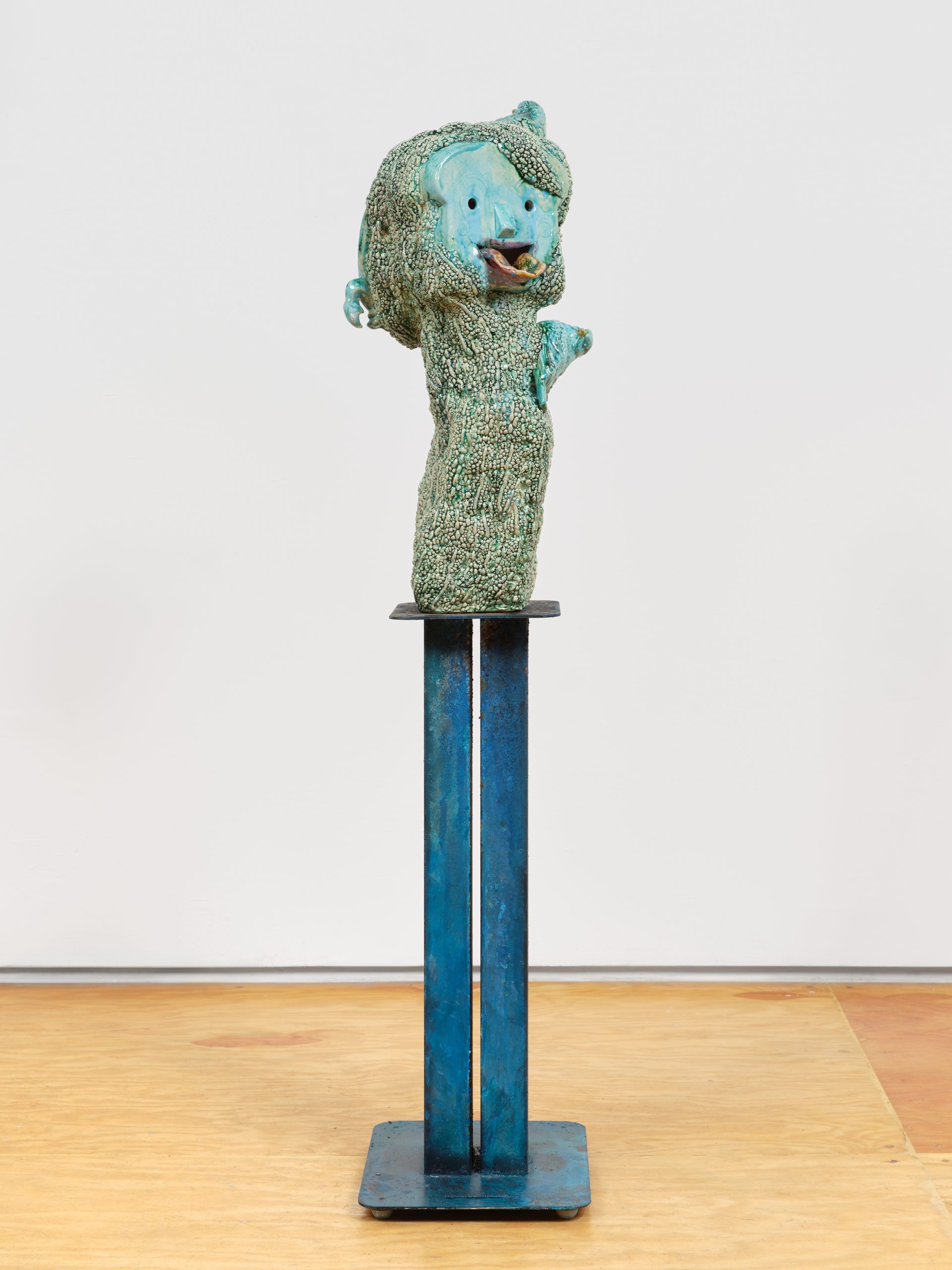Turquoise-glazed ceramic figure with a doll-like face, textured surface, and small bird on top, mounted on a tall blue metal pedestal.