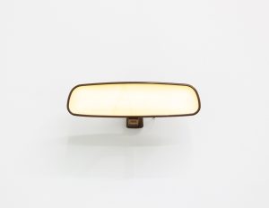 A minimalist wall-mounted sculpture resembling a car rearview mirror, centered against a white wall. The rounded rectangular form glows with soft warm light and is framed by a thin dark border, attached by a small central bracket. Its illumination casts a faint shadow below, giving the object a quiet, sculptural presence.