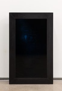 A dark, vertically oriented framed work leaning against a white wall on the floor. The wide black frame surrounds a nearly black reflective surface, within which a faint blue glow and barely visible shadowy form emerge near the center, giving the piece a quiet, enigmatic presence.