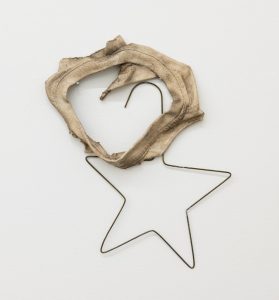 A wall-mounted mixed-media sculpture combining a bent metal wire hanger shaped like a five-pointed star with a worn, beige fabric form draped and stretched across its upper half. The fabric loop is frayed and stained, creating an irregular hollow center, while the exposed wire remains visible below, giving the piece a raw, improvised feel.