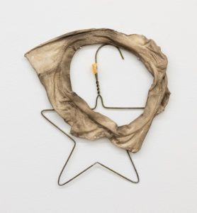 A wall-mounted sculpture made from a thin metal clothes hanger bent into a star-like shape and partially wrapped with a rough, beige fabric ring. The fabric looks worn, stiff, and frayed, with an uneven circular opening in the center, while the hanger’s hook and twisted neck remain visible above. The combination of soft textile residue and exposed wire gives the piece a fragile, handmade appearance.