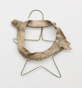 A wall-mounted sculpture made from a thin metal clothes hanger bent into a star-like shape and partially wrapped with a rough, beige fabric ring. The fabric looks worn, stiff, and frayed, with an uneven circular opening in the center, while the hanger’s hook and twisted neck remain visible above. The combination of soft textile residue and exposed wire gives the piece a fragile, handmade appearance.